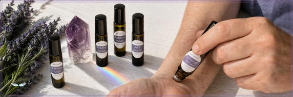 A person applies an essential oil drop from a small amber roller bottle onto their wrist. Several similar bottles, lavender sprigs, and a purple crystal are arranged on a light surface with a rainbow reflection.