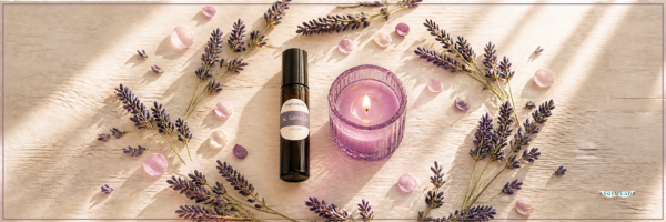 A small amger essential oil roller bottle and a lit purple glass candle are arranged on a light fabric surface with dried lavender sprigs, pink crystals, and soft window-light shadows.